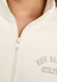 Felpa crema con zip, caratterizzata da un colletto rialzato e una grafica sottile "New Balance Athletics" in marrone vicino alla zip. Tessuto morbido e strutturato.