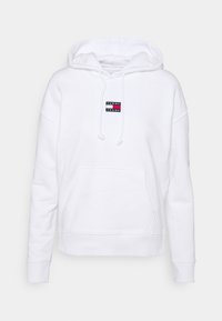 Sudadera blanca hecha de tela suave, que presenta un bolsillo tipo canguro en la parte frontal, cordones en la capucha y un pequeño parche con el logo en el pecho.