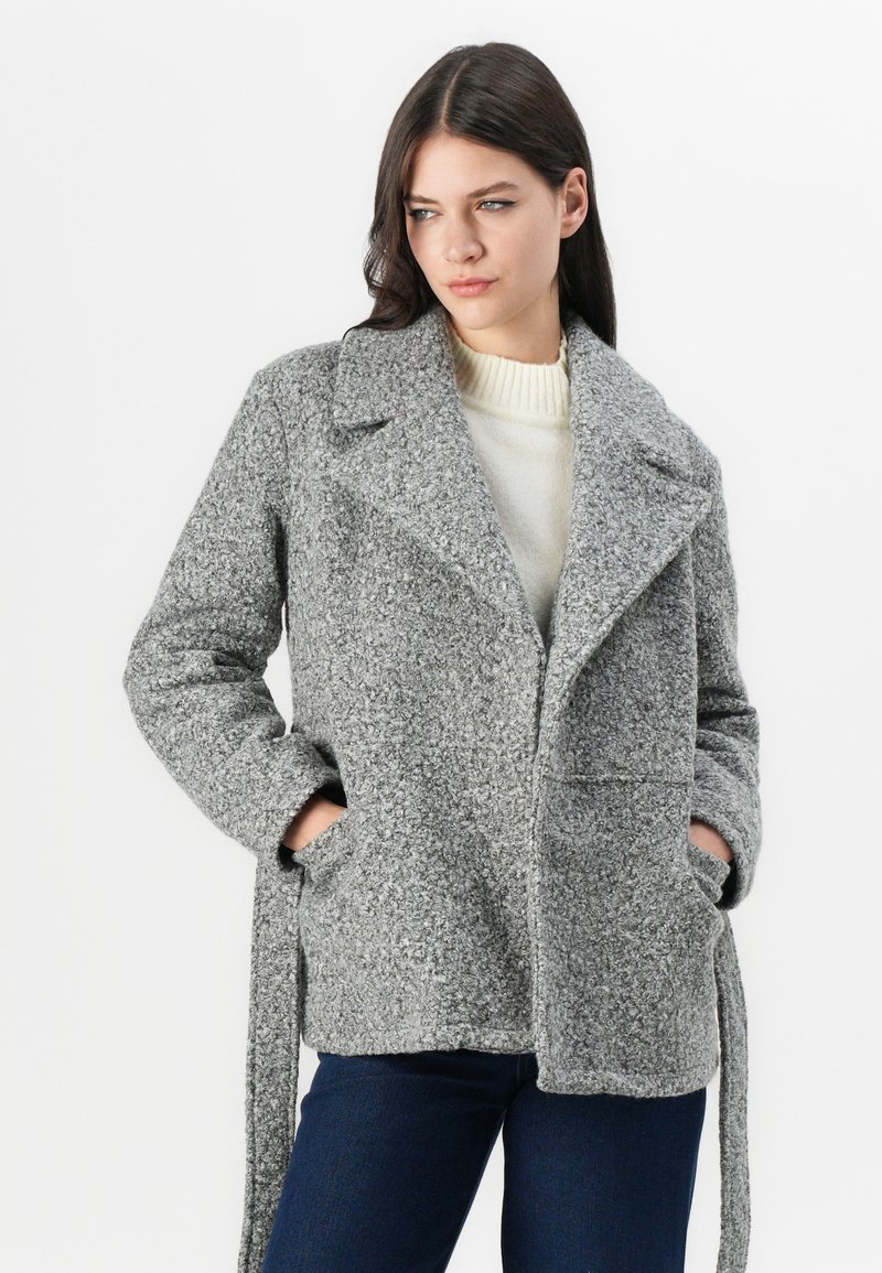 Manteau en mélange de laine gris avec une finition texturée, grands reverse, deux poches avant et un lien de ceinture. Porté sur un pull léger.