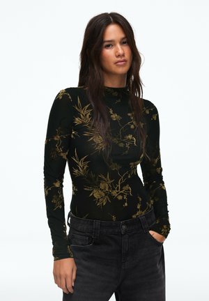 Femme aux longs cheveux foncés porte un haut noir à manches longues avec un imprimé floral doré et un jean foncé, debout devant un fond blanc uni.