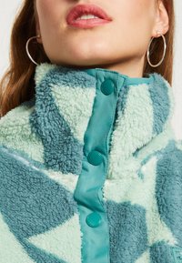 Fleecejack met geometrisch mintgroen en teal ontwerp. Heeft een hoge kraag, grote knopen en een fluffy textuur.