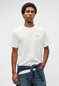 Λευκό κοντομάνικο μπλουζάκι με μικρό, σκούρο λογότυπο στο αριστερό στήθος. Συνδυασμένο με μπλε τζιν παντελόνι και ναυτικό πλεκτό πουλόβερ δεμένο γύρω από τη μέση.