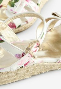 Kwiatowe sandały na koturnie z białymi paskami ozdobionymi różowymi i fioletowymi kwiatami, złotym zapięciem i plecioną jutatą.