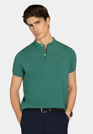 Mężczyzna ubrany w zieloną koszulkę Henley z krótkim rękawem i guzikami, granatowe spodnie, czarny pleciony pasek oraz zegarek na nadgarstek z jasnym paskiem.