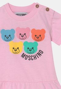Różowa bawełniana koszulka z pięcioma kolorowymi, bajkowymi twarzami misiów w kolorach niebieskim, różowym, pomarańczowym, żółtym i zielonym, z nazwą marki "Moschino" poniżej.