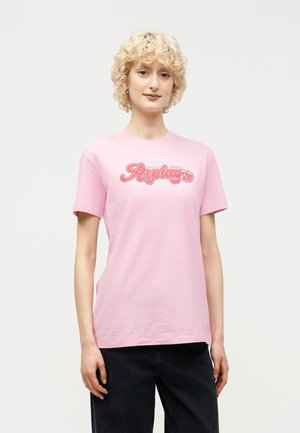 Młoda osoba o kręconych blond włosach, ubrana w pastelowo-różową koszulkę z logo "Replay" oraz czarne spodnie, na tle jednolitego jasnego tła.