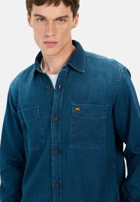 Granatowa koszula z denimu z kołnierzykiem zapinanym na guziki, dwoma przednimi kieszeniami i małym żółtym logo. Materiał wydaje się miękki i ma strukturalną powierzchnię.