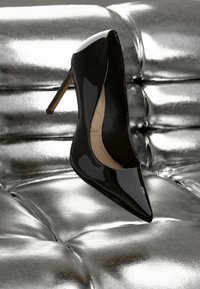 Escarpin noir en cuir verni à talon haut, avec un bout pointu et un talon aiguillé mince, sur un fond argenté brillant.