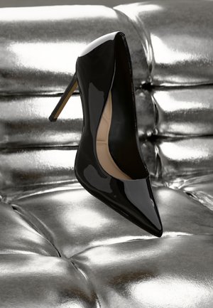 Musta lakattu nahkainen korkokenkä teräväkärkisellä muotoilulla ja hoikalla stiletto-korkolla, asetettuna kiiltävän hopean taustalle.