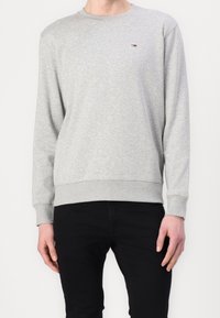 Jasnoszara, długorękawowa bluza z okrągłym dekoltem z małym prostokątnym logo na lewej piersi, noszona z czarnymi spodniami.