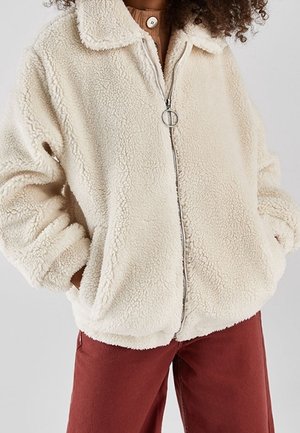 Person iført en cremefarvet sherpa lynlåsjakke med en rund lynlåstrækker og rustfarvede bukser, hænderne i jakkelommerne.