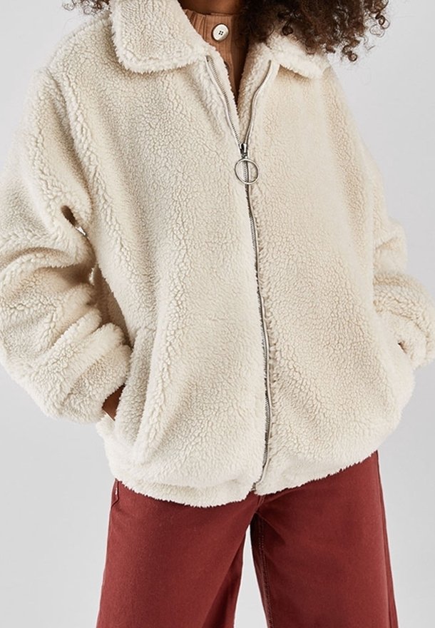 Personne portant une veste sherpa crème zippée avec une tirette de fermeture éclair ronde et un pantalon couleur rouille, les mains dans les poches de la veste.