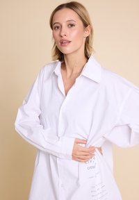 Hvit, oversized skjorte i bomull med klassisk krage og trykt tekstdetalj på siden, myk tekstur.