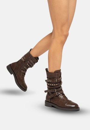 Botas de tobillo de cuero marrón con hebillas, tachuelas y correas. La suela robusta y los acentos texturizados realzan el diseño robusto.