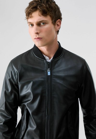 Jeune homme aux cheveux courts et bouclés, portant une veste en cuir noire zippée par-dessus une chemise blanche, sur un fond clair uni.
