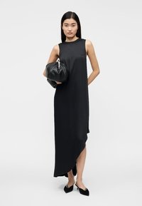 Vestido preto sem mangas com bainha assimétrica, confeccionado em tecido liso. Modelo segura uma mala redonda preta. Sapatos pretos e pontiagudos.