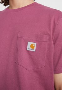 Kurzärmliges, runde-Hals-T-Shirt aus kastanienbraunem Baumwollstoff mit einer Brusttasche. Verfügt über ein quadratisches weißes Patch mit einem orangefarbenen Logo. Glatte Textur.
