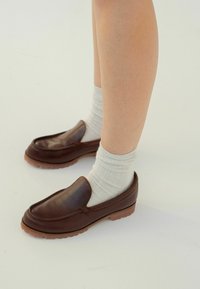 Scarpe slip-on in pelle marrone con texture liscia e suola in gomma, abbinate a calzini a coste grigio chiaro. Dettaglio di cucitura visibile sulle scarpe.
