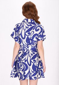 Blauw en wit gestippelde jurk met een kraag, korte mouwen en een ceintuur in de taille; de stof heeft een gestructureerde afwerking.