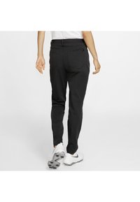 Person walking away wearing black pants with back pockets and white Nike golf shoes on a plain white background.