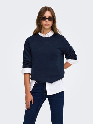 Jeune femme portant des lunettes de soleil foncées, un pull bleu marine sur une chemise blanche et un jean bleu foncé, debout avec une main sur la hanche.