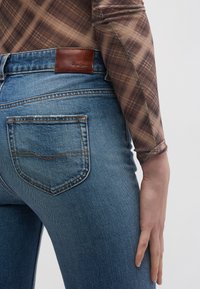 Εφαρμοστά μπλε τζιν με μεσαία πλύση, που διαθέτουν καφέ δερμάτινη ετικέτα, αντίθετη ραφή και κλασικό σχεδιασμό πίσω τσέπης.