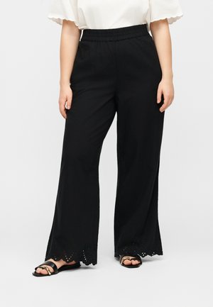 Kvinde iført sorte bukser med vide ben og bølgede hulmønstrede kanter, kombineret med sorte sandaler og en hvid kortærmet bluse.