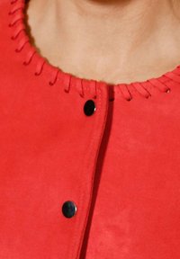 Vêtement rouge avec encolure ronde présentant un détail de couture en boucle et deux boutons-pression noirs sur la fermeture avant.