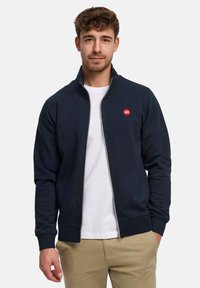 Sweatshirt zippé bleu marine en tissu doux avec un col montant, présentant un petit logo rouge sur la poitrine et des poignets côtelés. Porté sur un t-shirt blanc.