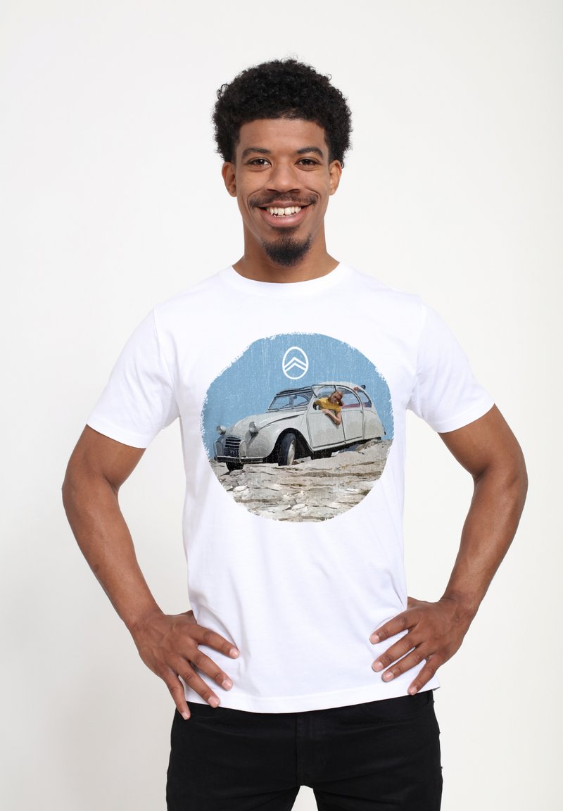 Hvid bomulds t-shirt med et trykt cirkulært grafisk billede af en vintage hvid bil med en person, der læner sig ud af vinduet, sat mod en blå baggrund.