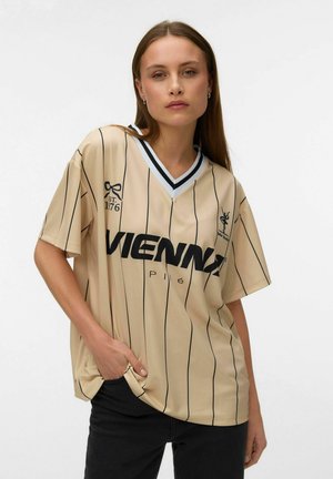 Jonge vrouw met lang bruin haar, gekleed in een beige oversized overhemd met krijtstreep en "VIENNA" gedrukt op de borst, poseert met haar hand in de zak.