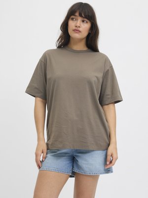 Woman with long dark hair wearing a loose brown t-shirt and light blue denim shorts standing against a plain white background.