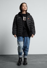 Giubbotto imbottito nero con trapuntatura orizzontale, cappuccio e dettagli del logo. Abbinato a una t-shirt grafica nera e jeans a patchwork blu chiaro. Stivali neri.