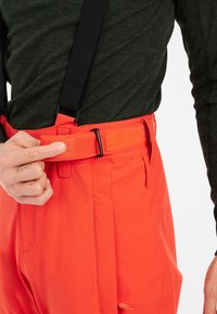 Pantalons orange vif avec bretelles ajustables et fermeture à taille en Velcro, fabriqués à partir d'un matériau lisse et durable. Poches latérales visibles.