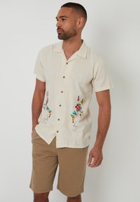 Hellbeige kurzärmliges Hemd mit buntem Blumenstickerei, mit durchgehender Knopfleiste und lockerem Schnitt. Getragen mit beigen Shorts.