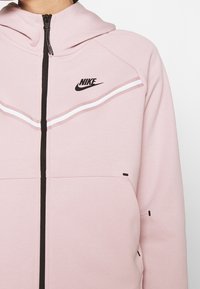 Felpa Nike rosa chiaro con zip frontale, hardware nero e strisce bianche sul petto. Presenta tasche laterali per la conservazione.