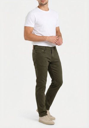 Homme portant un pantalon slim coupe olive, un t-shirt blanc à encolure ronde et des baskets beige, debout les mains jointes.
