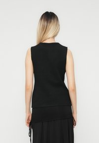 Kvinna med långt blont hår som bär en ärmlös svart topp och en svart kjol med snörningsdetaljer, stående mot en enkel ljus bakgrund.