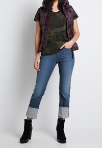 Vadderad lila väst över en kamouflage-t-shirt, kombinerad med ljusblå jeans med uppvikta muddar och svarta ankelkängor.