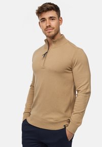 Pull-over zippé beige avec col montant, design ajusté, poignets et ourlet côtelés, fabriqué en tissu doux, associé à un pantalon navy.