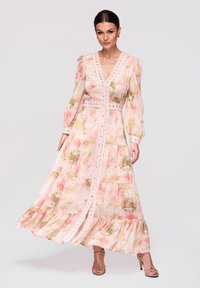 Femme debout portant une robe longue à manches longues à fleurs roses avec des bordures en dentelle, un décolleté en V, des détails à boutons, et des sandales à talons hauts roses.