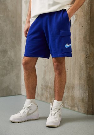 Mann trägt blaue Cargoshorts mit Nike-Logo und weiße hohe Nike-Sneaker, steht auf einem hellgrauen Boden vor einer Betonwand.