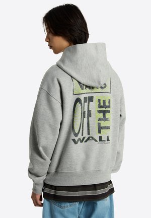 Persona con una sudadera gris de Vans con el logo "Vans Off The Wall" en negro y verde en la espalda, combinada con vaqueros azul claro y varias camisas superpuestas.