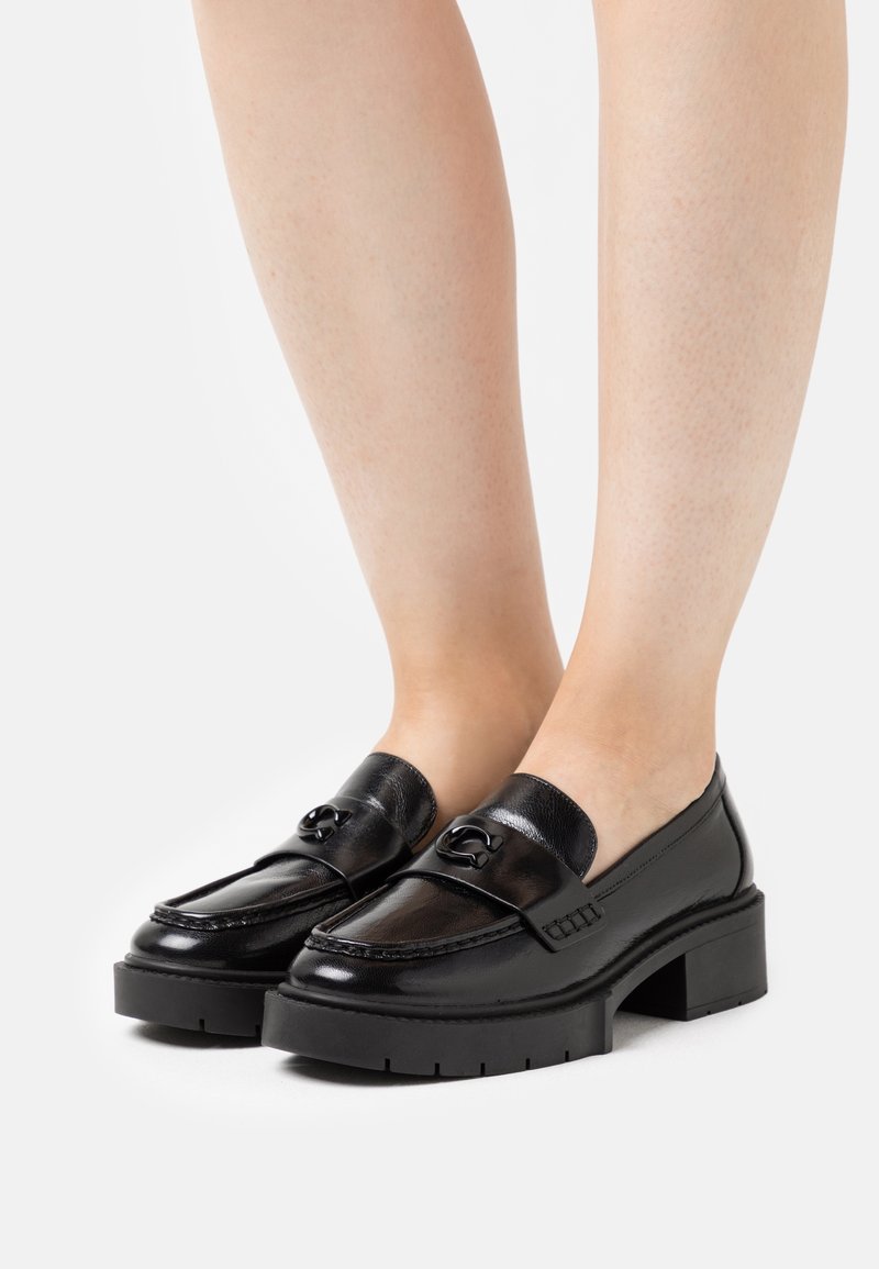 Mocassins noirs en cuir à talons épaissis avec boucle décorative portés pieds nus sur fond blanc.