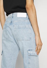 Pantalones cargo de denim azul claro con bolsillos en la parte trasera y lateral. Presenta una etiqueta de cinturón blanca lisa. Textura suave, diseño de pierna recta.