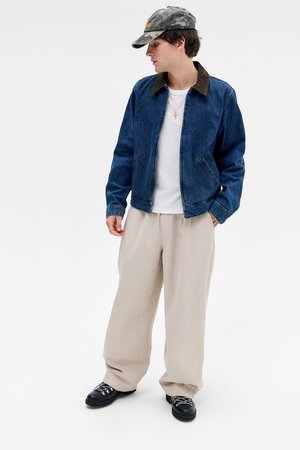 Ung mann iført denimjakke, hvit skjorte, beige vide bukser, svarte sko og kamuflasje baseballcap som står mot en hvit bakgrunn.