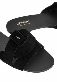 Czarne zamszowe sandały z krzyżowym panelem, gładką czarną podeszwą i małym złotym akcentem. Logo marki jest widoczne na wkładce.