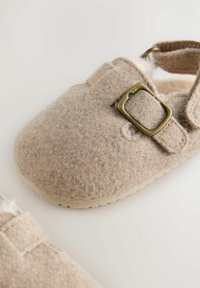 Chaussons en feutre beige avec une forme arrondie, dotés d'une sangle à boucle, d'un intérieur doux et d'une semelle en caoutchouc de couleur claire.