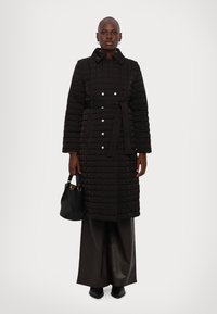 Melna piegriezta gara mētelis ar jostu viduklī, sudraba pogām un apkakli. Uzvilkts virs tumšiem platām kājām biksēm un turēt melnu rokassomu.