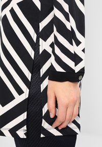 Langärmliges Oberteil in Schwarz-Weiß mit Muster, das diagonale Streifen zeigt, Belüftungsdetails an den Seiten und einem Knopfmanschette. Weicher Stofftextur.